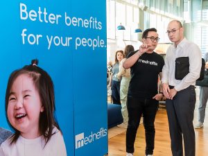 "Better benefits for your people" written on blue backdrop with a photo of a smiling child on the left and the Mednefits logo on the bottom right corner. Next to the backdrop are two men talking. | Photo by Mednefits/NHA File Photo