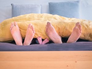 Three people lying in bed with a yellow blanket. Visible in the photo are only the three pairs of feet of a mother, a baby and a father. | Photo for illustrative purposes only. | Photo by Simon Berger/Unsplash/NHA File Photo