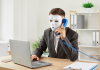 Anonymous man disguised in a full-face white mask while sitting at an office desk with a laptop and holding a phone receiver, making a phone call, trying to scam people and steal their money through fake job ads. | Photo by studioroman via Canva Pro / NHA File Photo