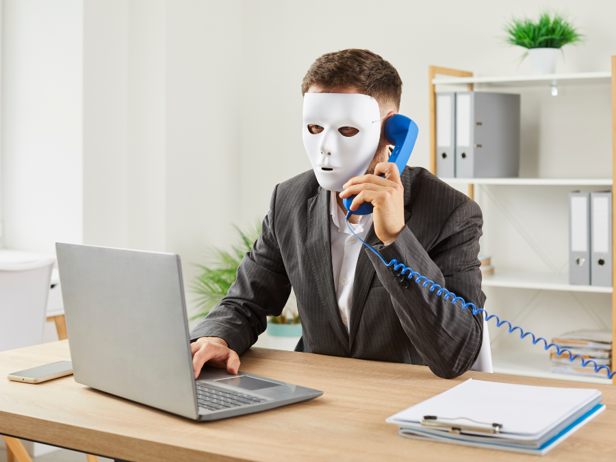 Anonymous man disguised in a full-face white mask while sitting at an office desk with a laptop and holding a phone receiver, making a phone call, trying to scam people and steal their money through fake job ads. | Photo by studioroman via Canva Pro / NHA File Photo